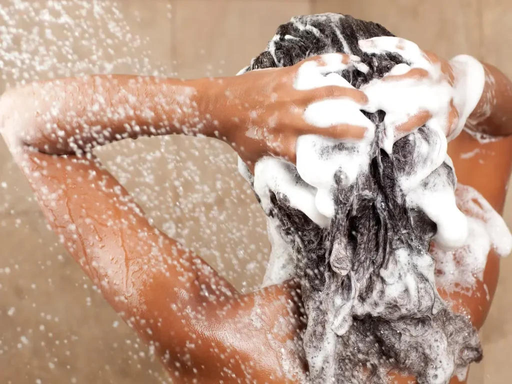 A black woman washing her hair with lathered up shampoo in the shower