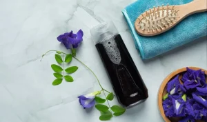 A clear bottle of dark coloured shampoo on a surface with a hair brush and flowers beside it